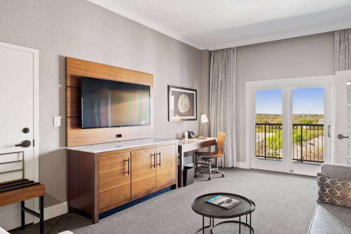 A modern hotel room with a large TV, a desk with a chair, a sofa, and a balcony with a view.