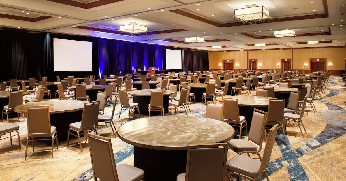 A large conference room with multiple round tables and chairs, a stage with a screen, and a carpeted floor.