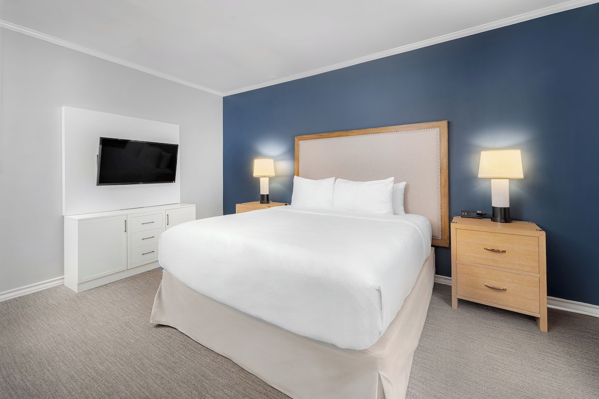 A neatly arranged hotel room with a large bed, two bedside tables with lamps, a flat-screen TV mounted on the wall, and a small cabinet.