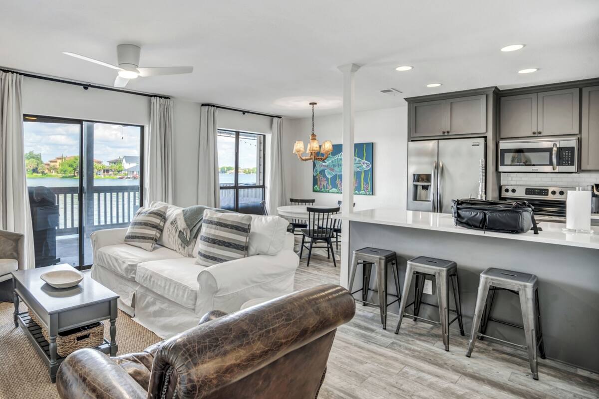 A modern living room with a sofa, armchair, and a dining area with a table and chairs. The kitchen features stainless steel appliances and a bar area with stools.