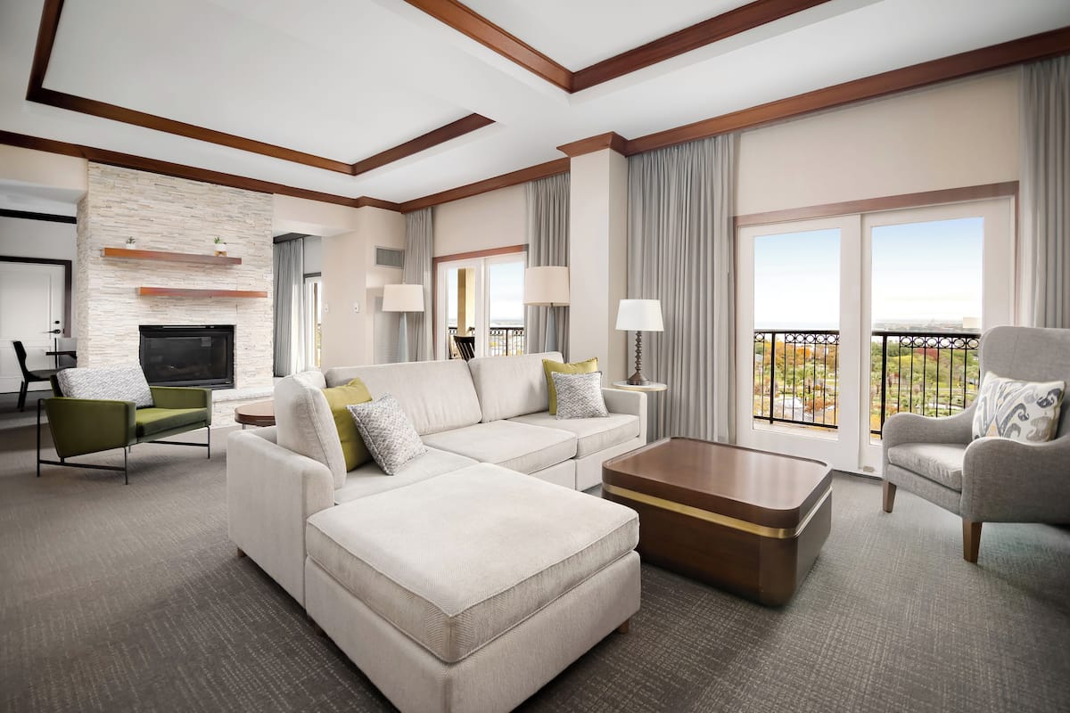 A modern hotel room with a sectional sofa, a flat-screen TV, a fireplace, a balcony with a view, and a dining area.