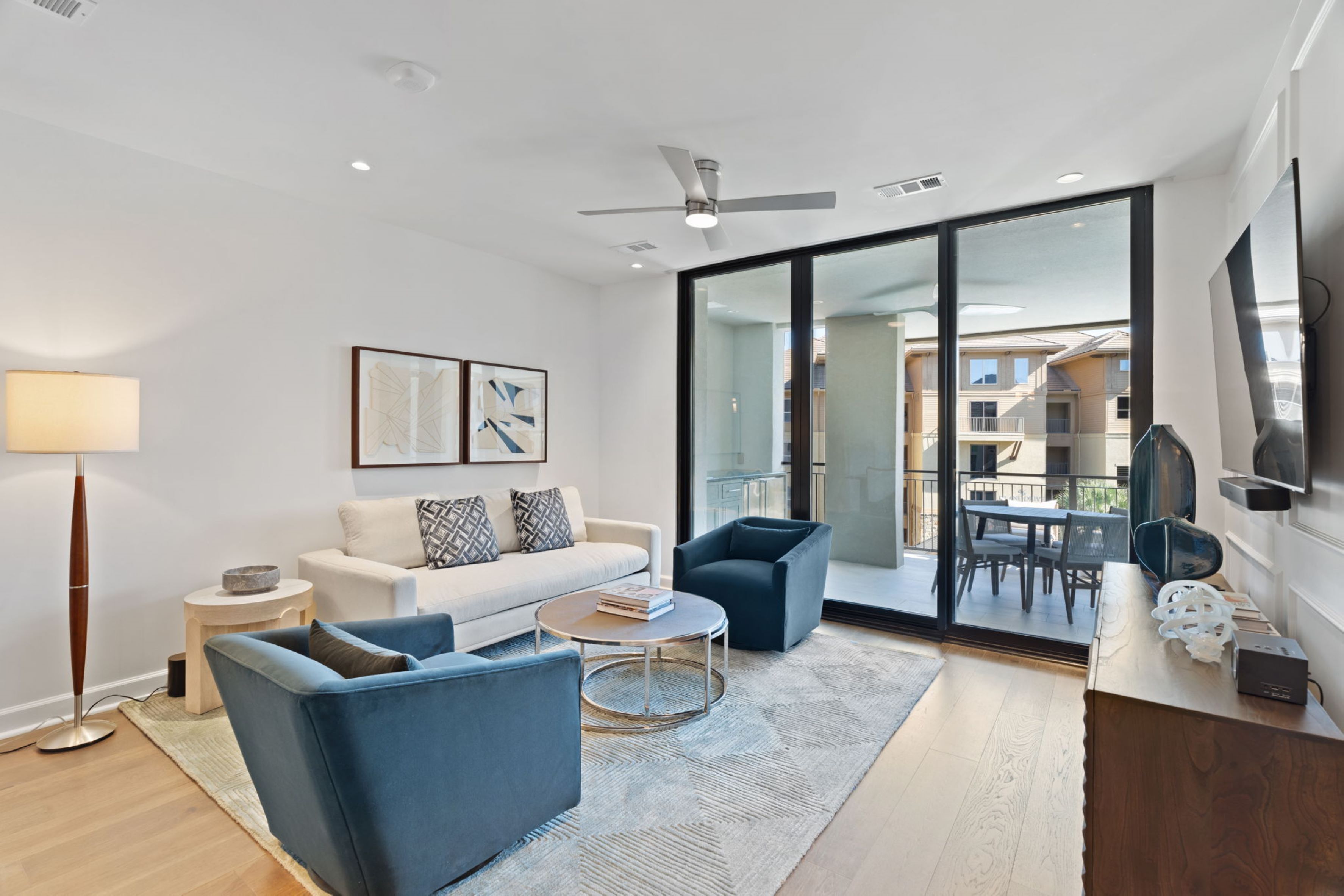 Sala de estar moderna com sofá branco, poltronas azuis, mesa de centro e abajur. Há uma janela grande com vista para uma varanda e edifícios.