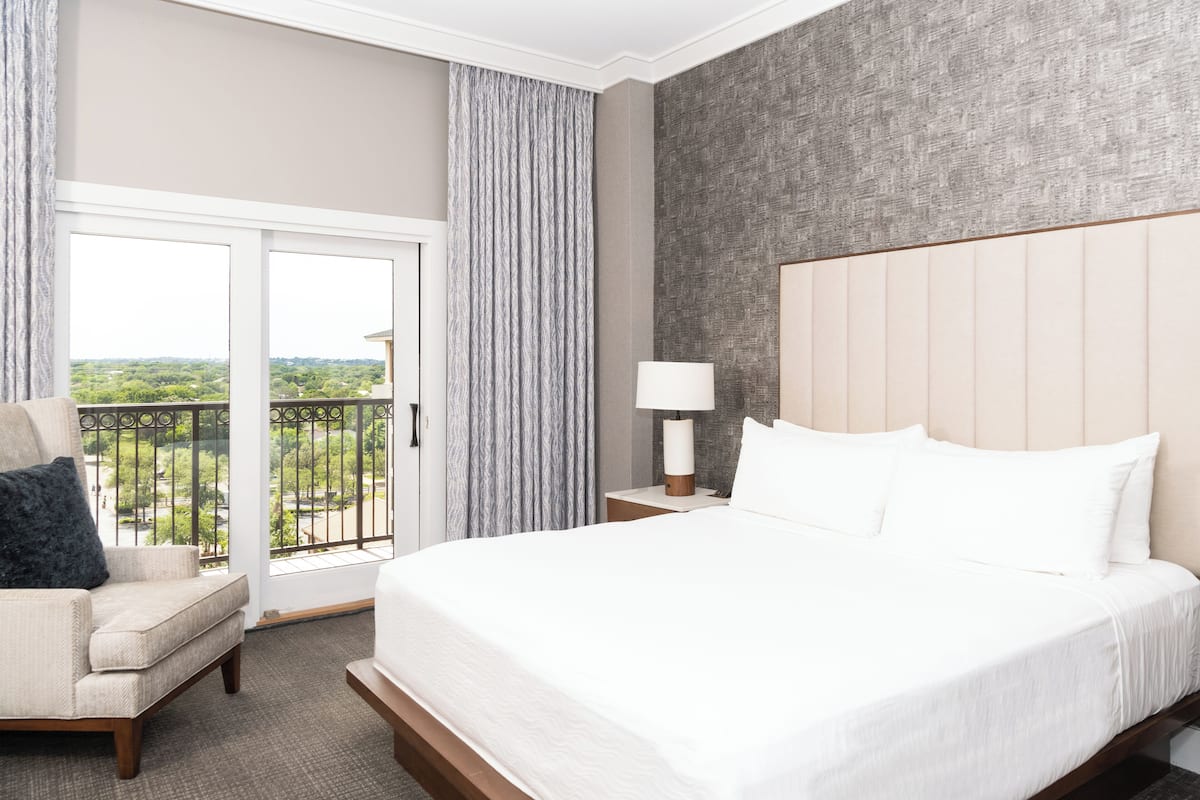 A hotel room with a large bed, a chair, a nightstand, a lamp, and a view of greenery through the window.