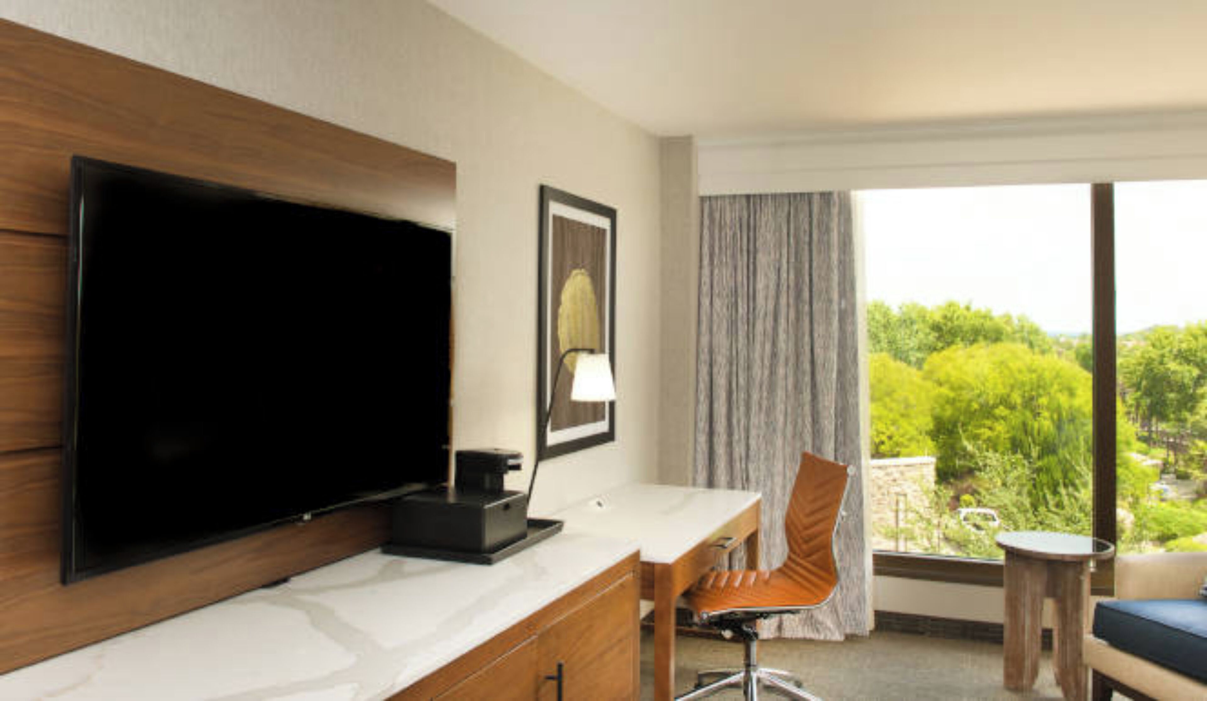 A hotel room with a flat-screen TV, a desk with a lamp, a chair, and a view of greenery outside.
