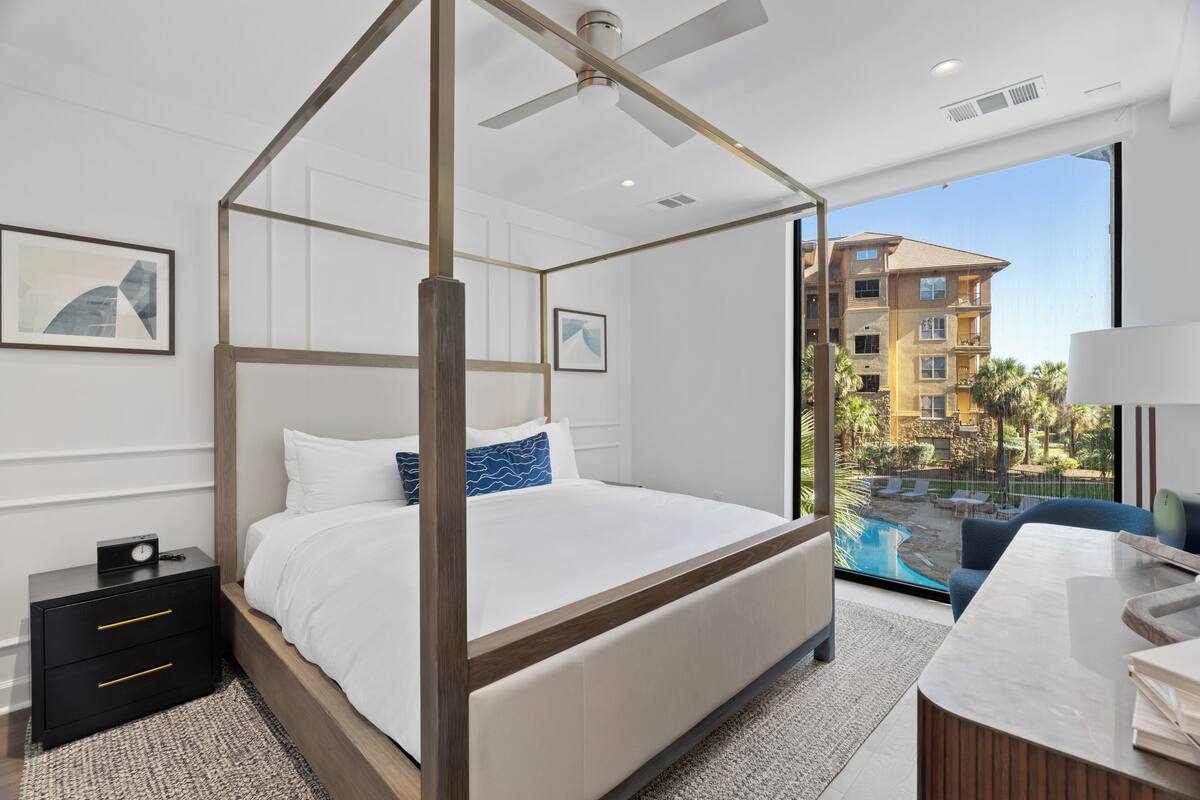 A modern hotel room with a four-poster bed, a desk with a lamp, and a view of a pool and buildings.