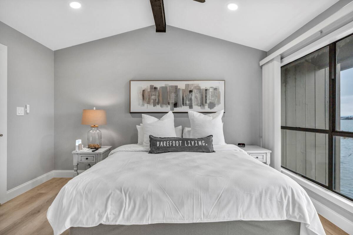 A bedroom with a large bed, a nightstand with a lamp, a framed artwork on the wall, and a window with a view of the water.