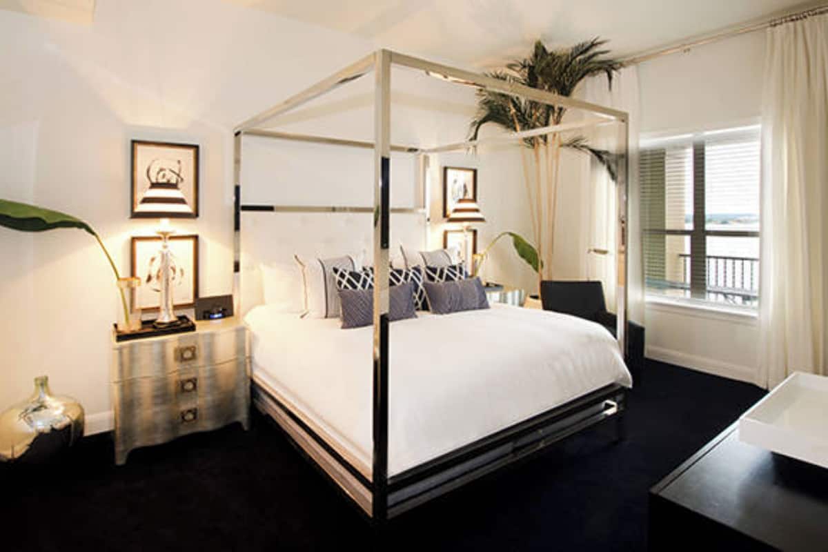 A four-poster canopy bed with a white bedspread, bedside tables with lamps, framed artwork on the walls, and a view of the outdoors through the window.