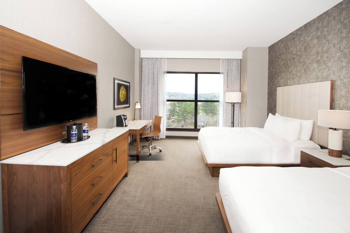 A hotel room with a large flat-screen TV, a desk with a chair, two beds, and a large window with a view of trees.
