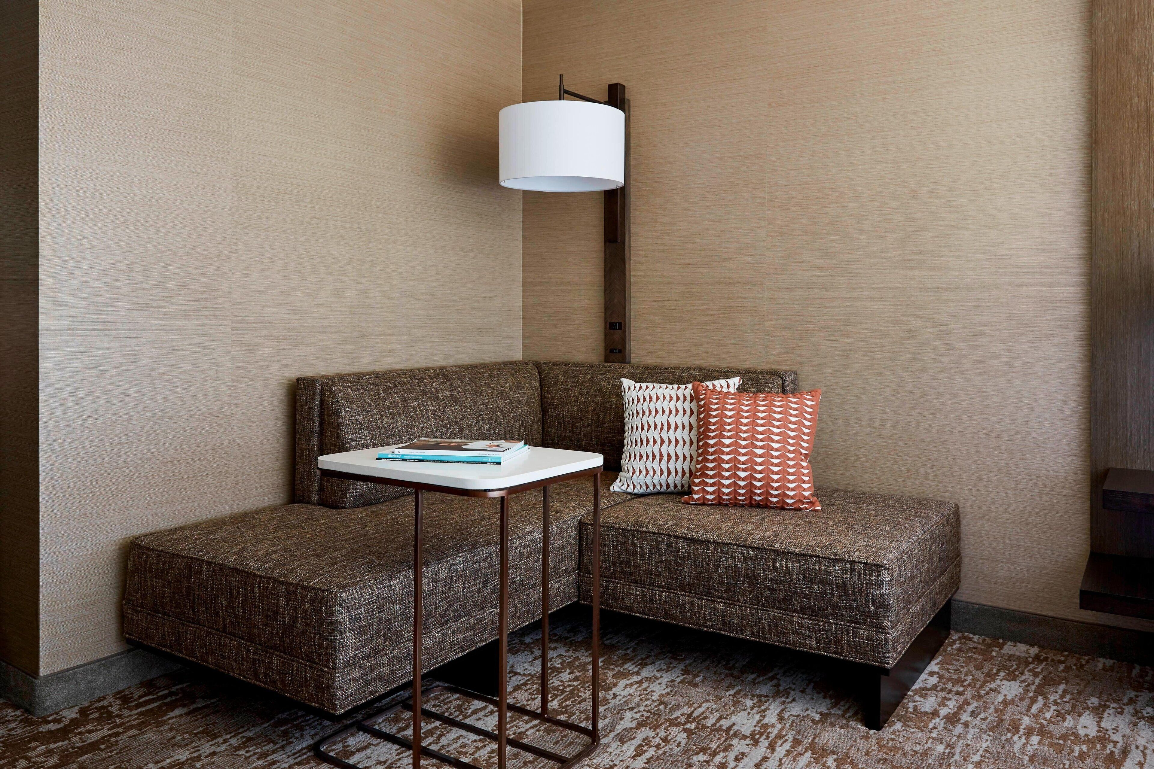 A corner seating area with a brown upholstered sofa, a small white side table, a patterned cushion, and a white lamp.