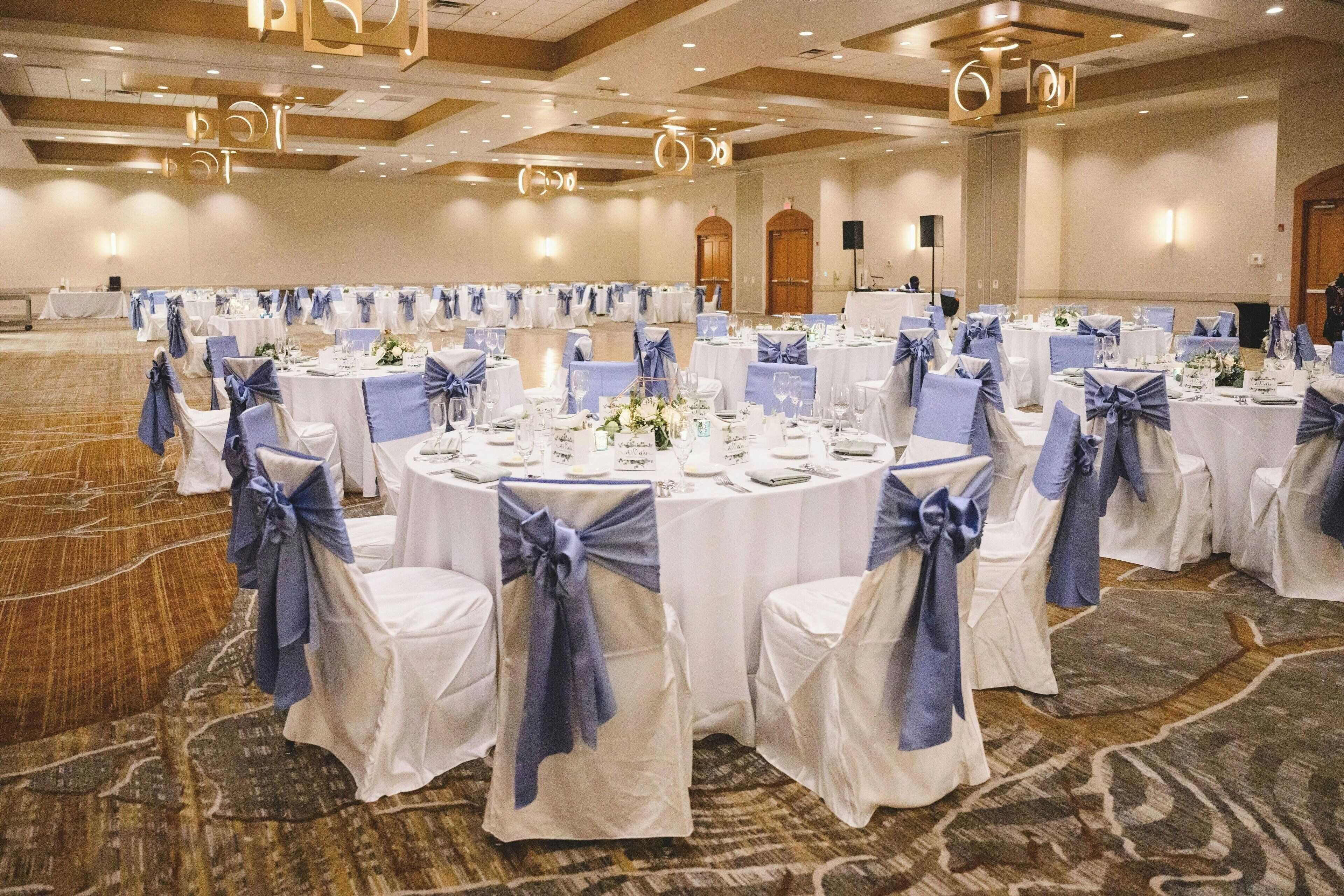 A banquet hall with round tables set for a formal event, featuring white tablecloths and blue chair covers.