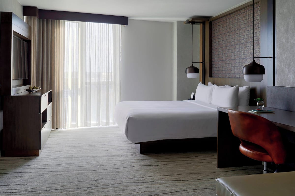 A hotel room with a large bed, a desk with a chair, a TV, and a large window with curtains.