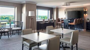 Habitación de hotel con comedor, bar y televisión. Hay varias sillas y mesas, además de un ventanal con vistas al exterior.