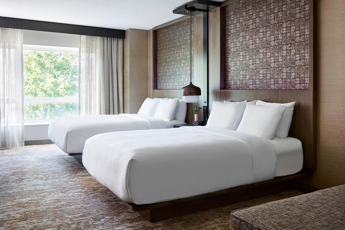 A hotel room with two beds, a large window with curtains, a wall with a textured pattern, and a hanging light fixture.