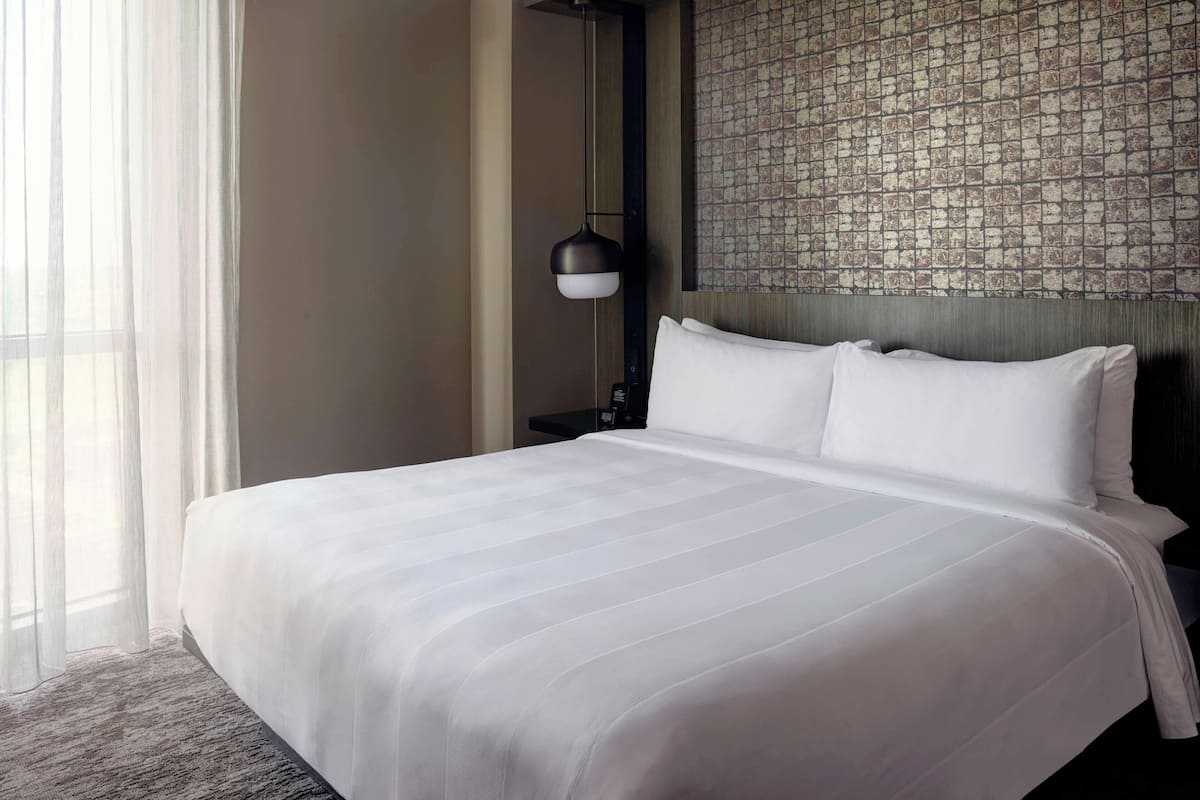 A neatly made bed with white linens, a wall with a patterned wallpaper, and a floor lamp.