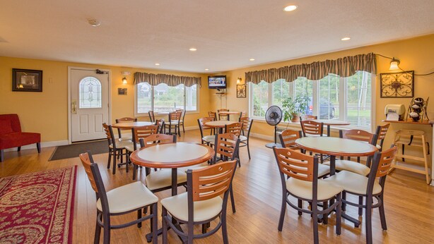 A dining area with multiple round tables and chairs, a television mounted on the wall, and large windows with curtains. - Coachman Inn (Kittery)