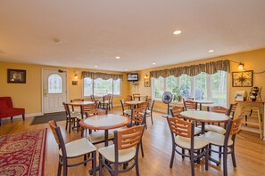 A dining area with multiple round tables and chairs, a television mounted on the wall, and large windows with curtains.