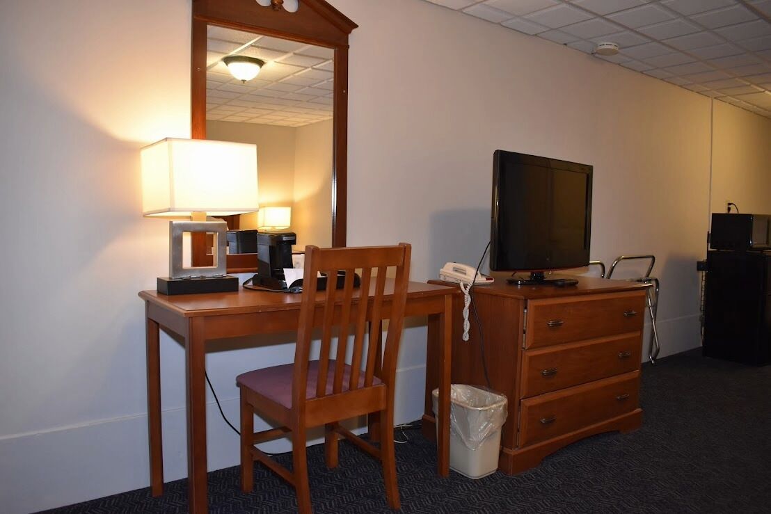 A hotel room with a wooden desk, a chair, a television, a telephone, a mirror, and a dresser.