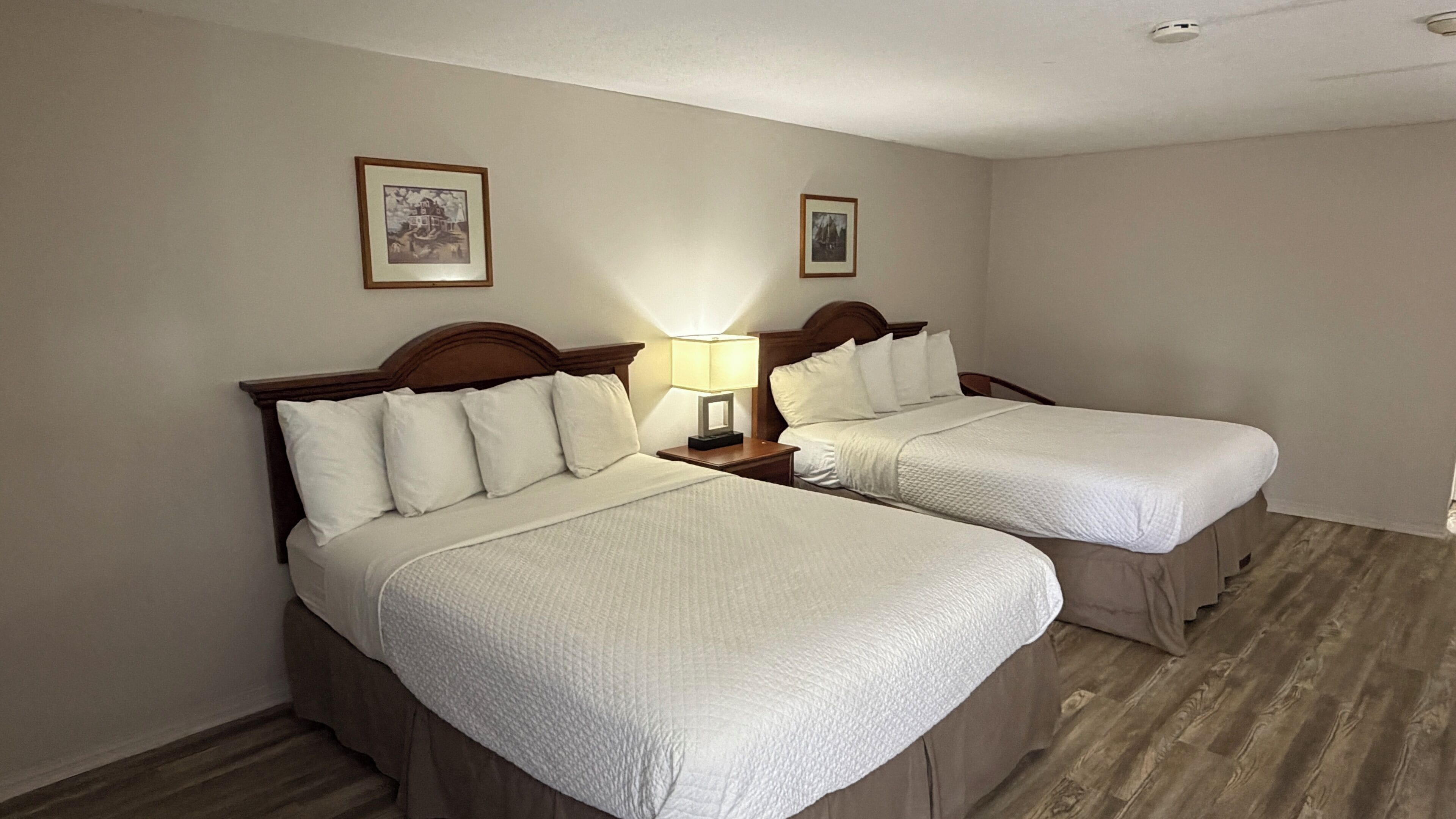 A hotel room with two beds, a nightstand with a lamp, and two framed pictures on the wall.
