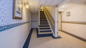 A hallway with patterned wallpaper, a carpeted floor, a staircase with a handrail, and a framed picture on the wall. - Coachman Inn (Kittery)