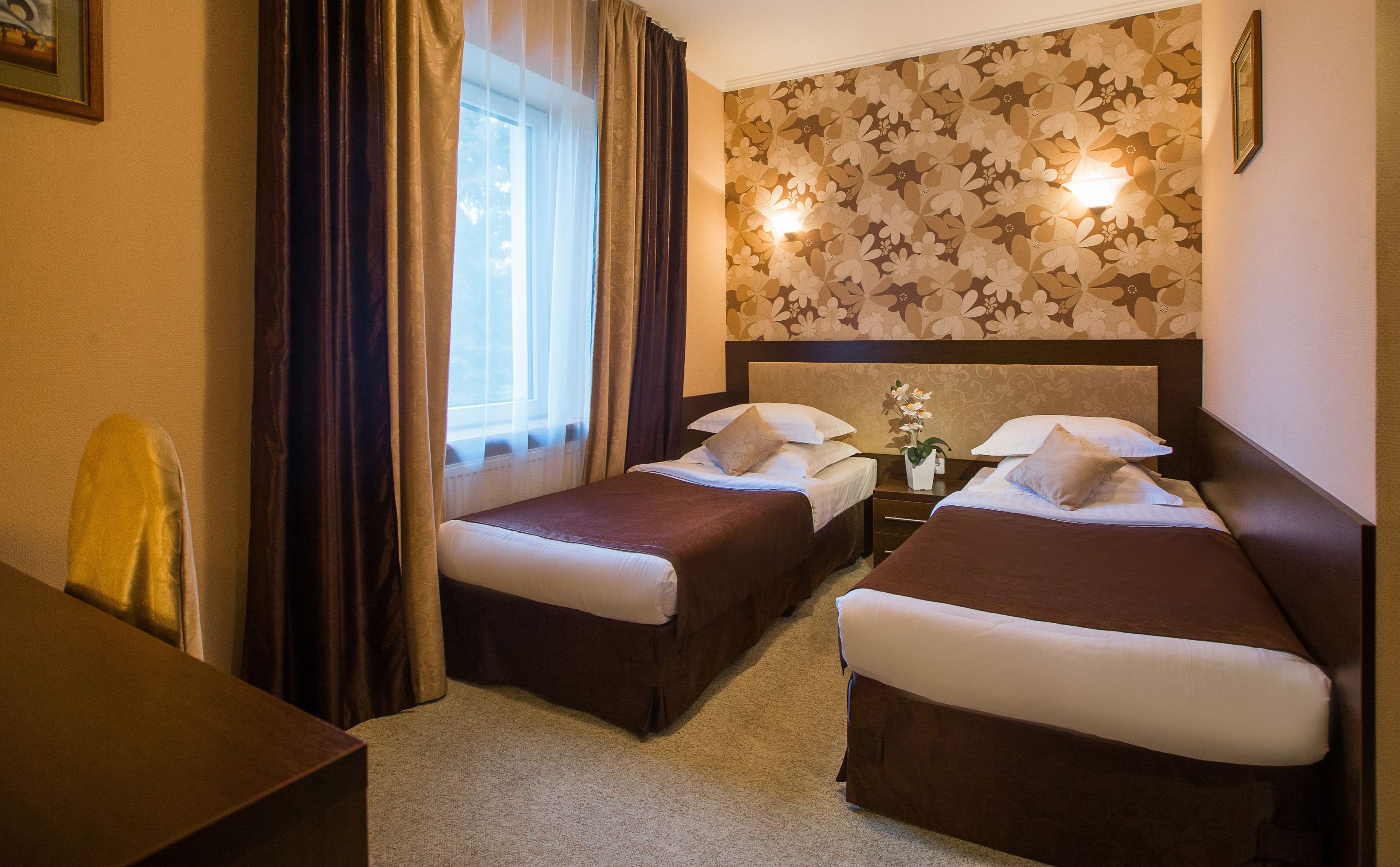 A hotel room with two beds, a desk, a chair, a window with curtains, and a floral wallpaper.