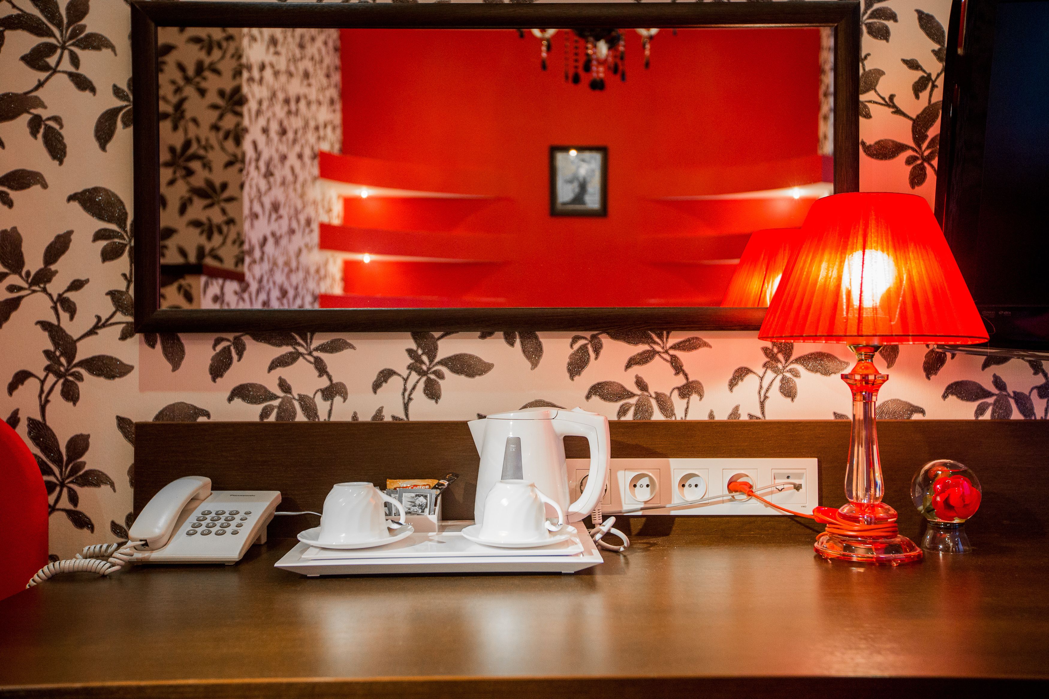 A hotel room with a red wall, a mirror, a red lamp, a telephone, a coffee maker, and a small decorative item.