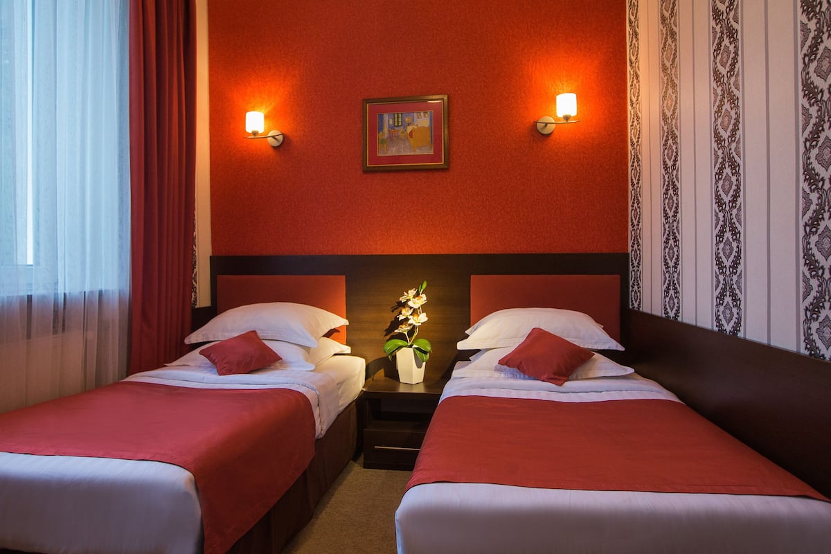 A hotel room with two beds, red bedding, a nightstand with a vase of flowers, and two wall-mounted lamps.