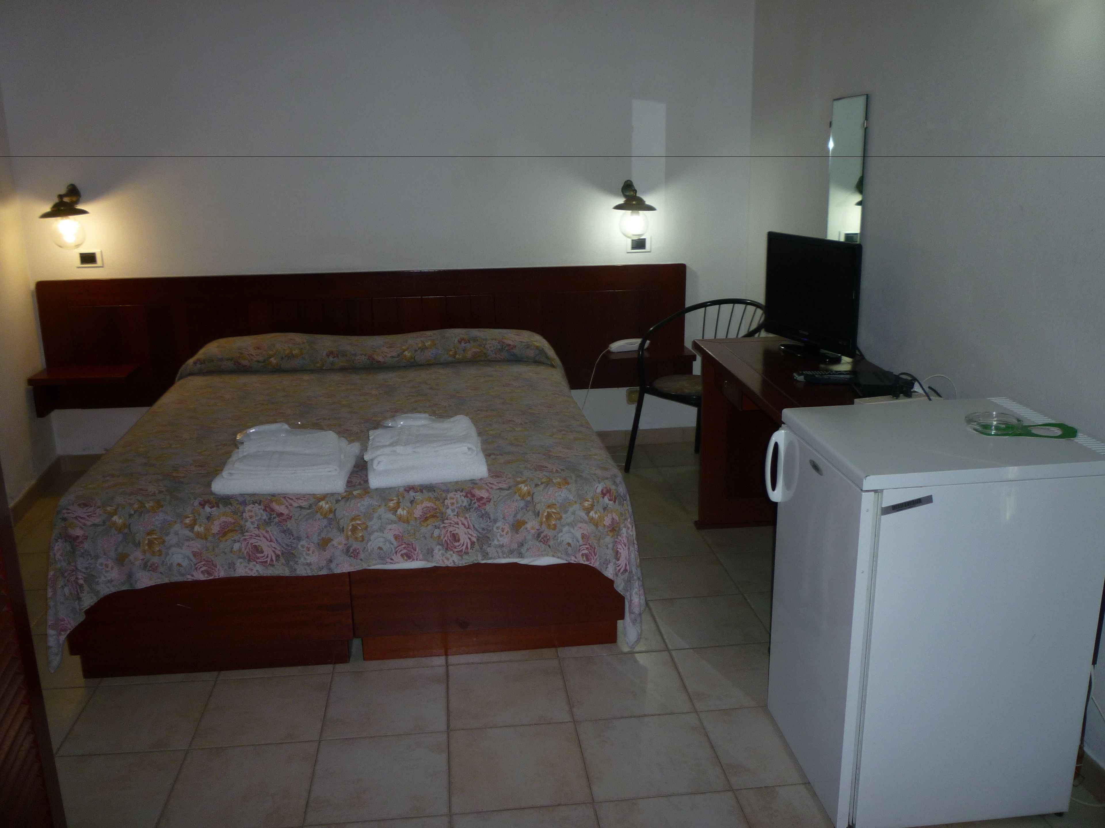 A hotel room with a bed, a desk with a television, a chair, and a small refrigerator.