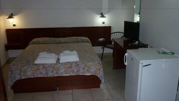 A hotel room with a bed, a desk with a television, a chair, and a small refrigerator.