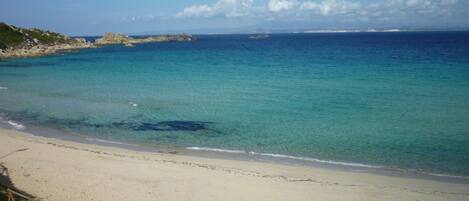 A beach with clear turquoise water, sandy shore, and rocky outcrops in the distance.