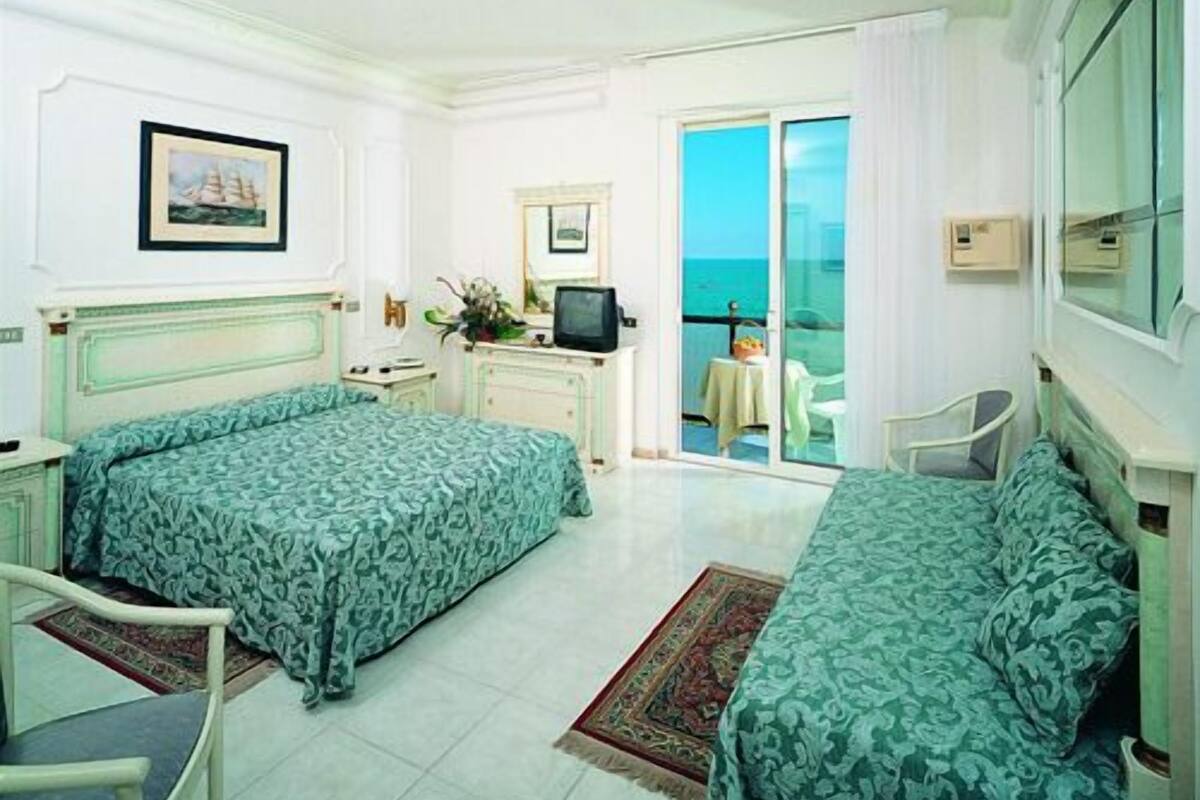 A hotel room with two beds, a desk, a chair, a television, and a balcony with a view of the sea.
