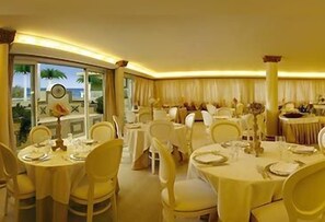 A banquet hall with round tables, white chairs, and a view of palm trees through large windows. - Grand Hotel Michelacci (Gabicce Mare)
