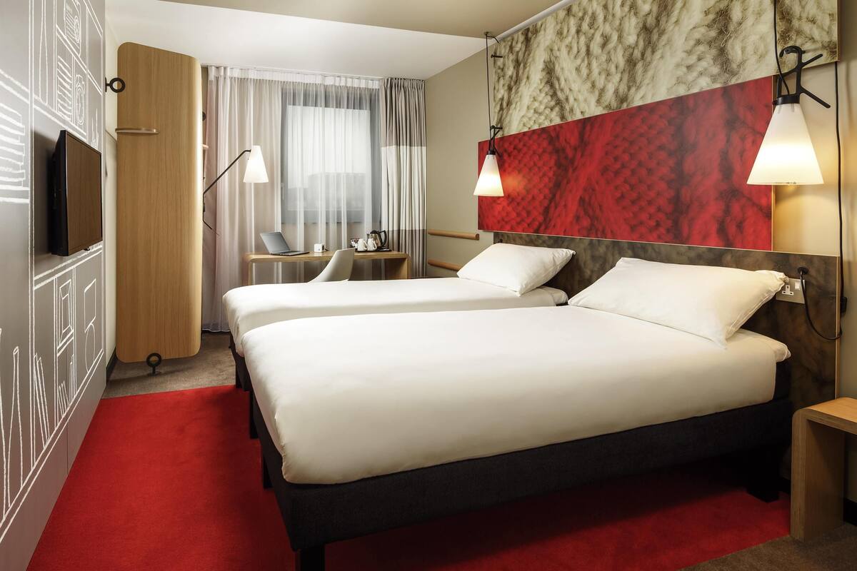 A hotel room with two beds, a desk with a laptop, a TV, and a red carpet.