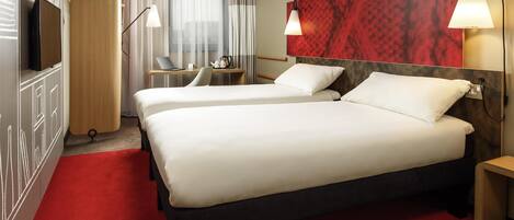 A hotel room with two beds, a desk with a laptop, a TV, and a red carpet.