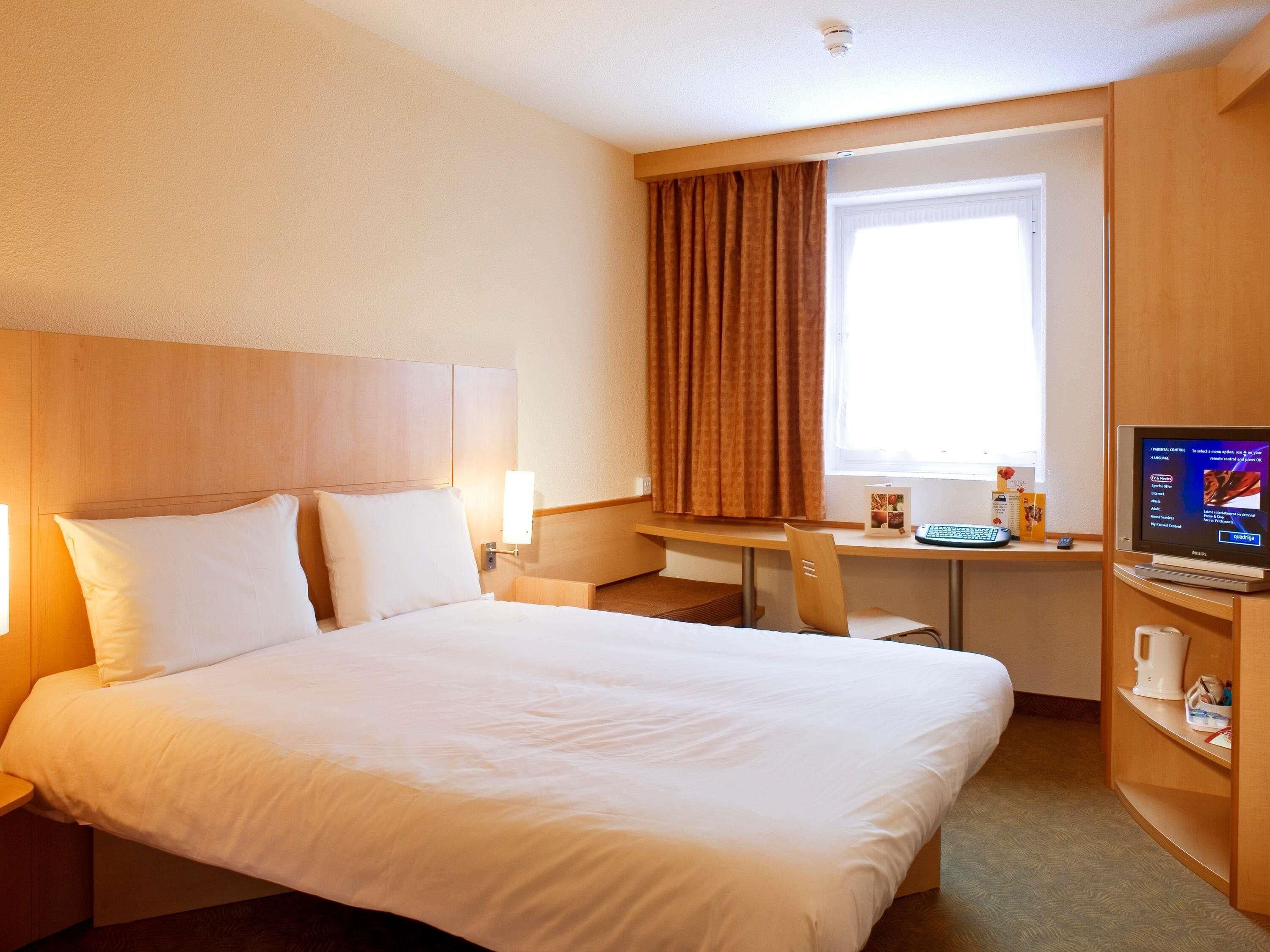 A hotel room with a large bed, a desk with a computer, a coffee maker, and a window with curtains.