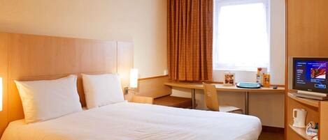 A hotel room with a large bed, a desk with a computer, a coffee maker, and a window with curtains.