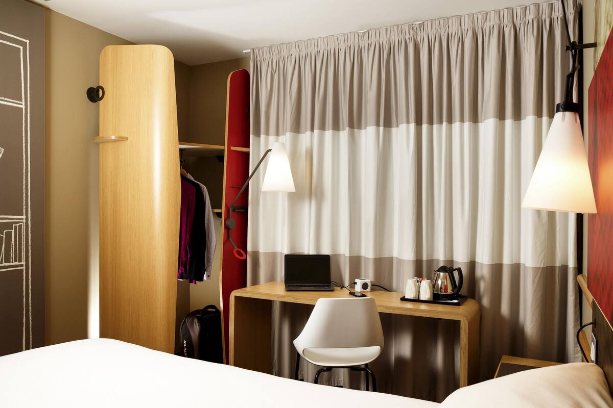 A hotel room with a bed, a desk with a laptop, a chair, a wardrobe, and a curtain.