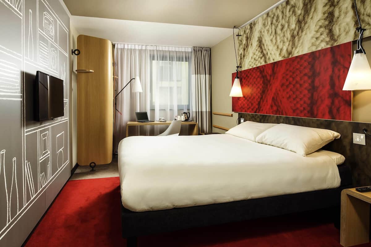 A hotel room with a bed, a desk with a laptop, a TV, and a red carpet.
