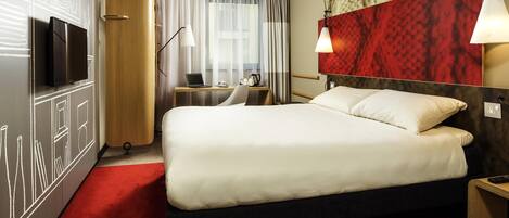 Ein Hotelzimmer mit einem Bett, einem Schreibtisch mit Laptop, einem Fernseher und einem roten Teppich.
