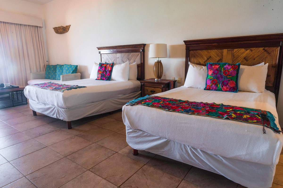 Habitación de hotel con dos camas, cada una con colcha blanca y almohadas coloridas. Hay una mesita de noche con lámpara entre las camas, y una ventana con cortinas al lado izquierdo.