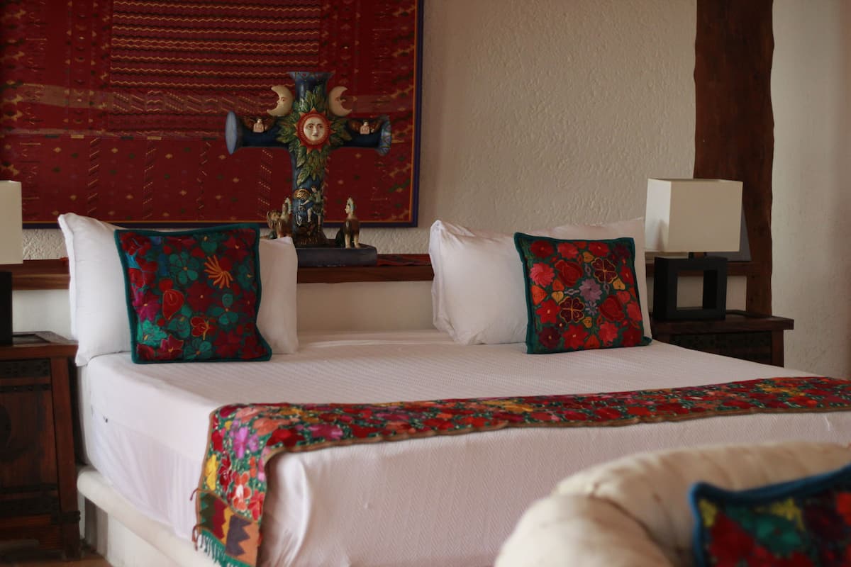 Una cama bien hecha con almohadones florales, una cruz decorativa y un tapiz rojo en la pared.