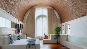 A bedroom with a brick ceiling, a large bed with white bedding, a wooden floor, a window with curtains, and a plant. - Casa de los Sueños Boutique Hotel (Isla Mujeres)