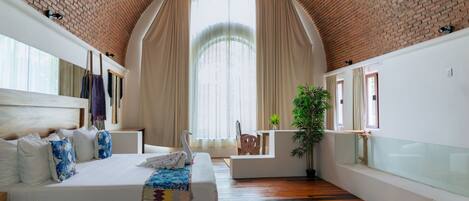 Een slaapkamer met een bakstenen plafond, een groot bed met wit beddengoed, een houten vloer, een raam met gordijnen en een plant.