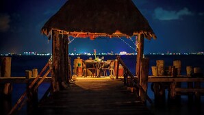 Une jetée en bois mène à une structure à toit de chaume, dotée de tables et de chaises, et d’où l’on peut admirer un plan d’eau avec les lumières de la ville en arrière-plan.