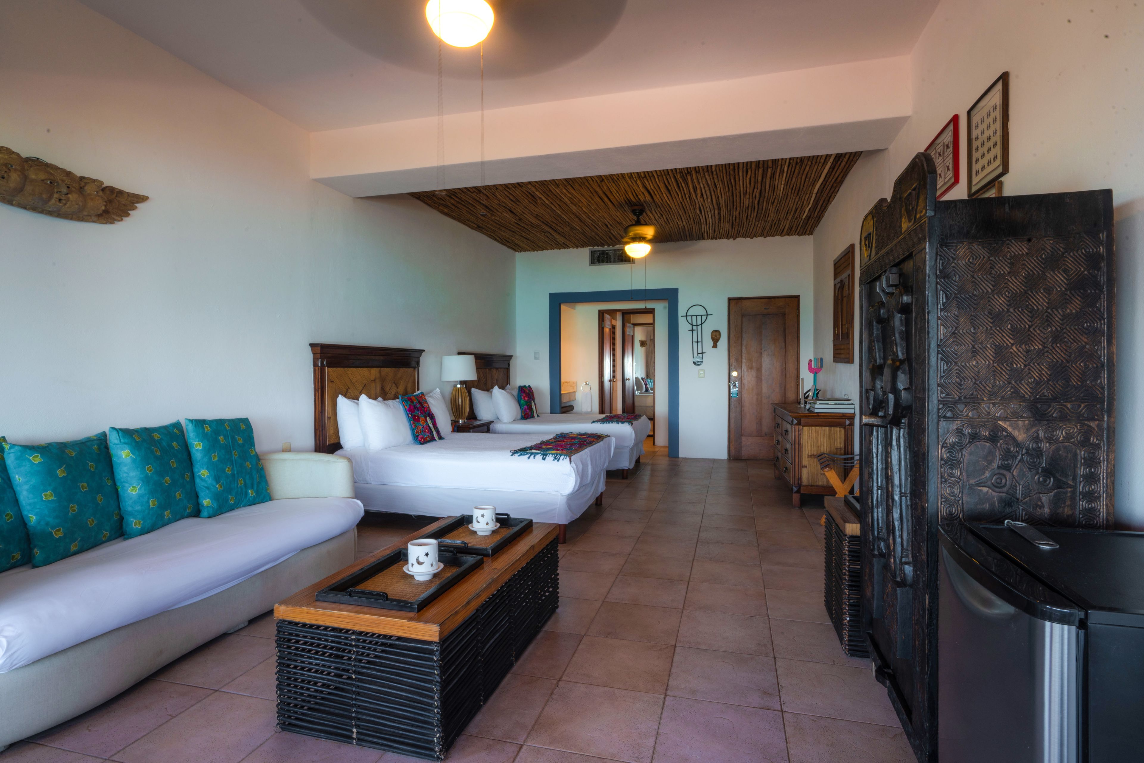 A hotel room with a sofa, two beds, a coffee table, a refrigerator, and a wooden ceiling.