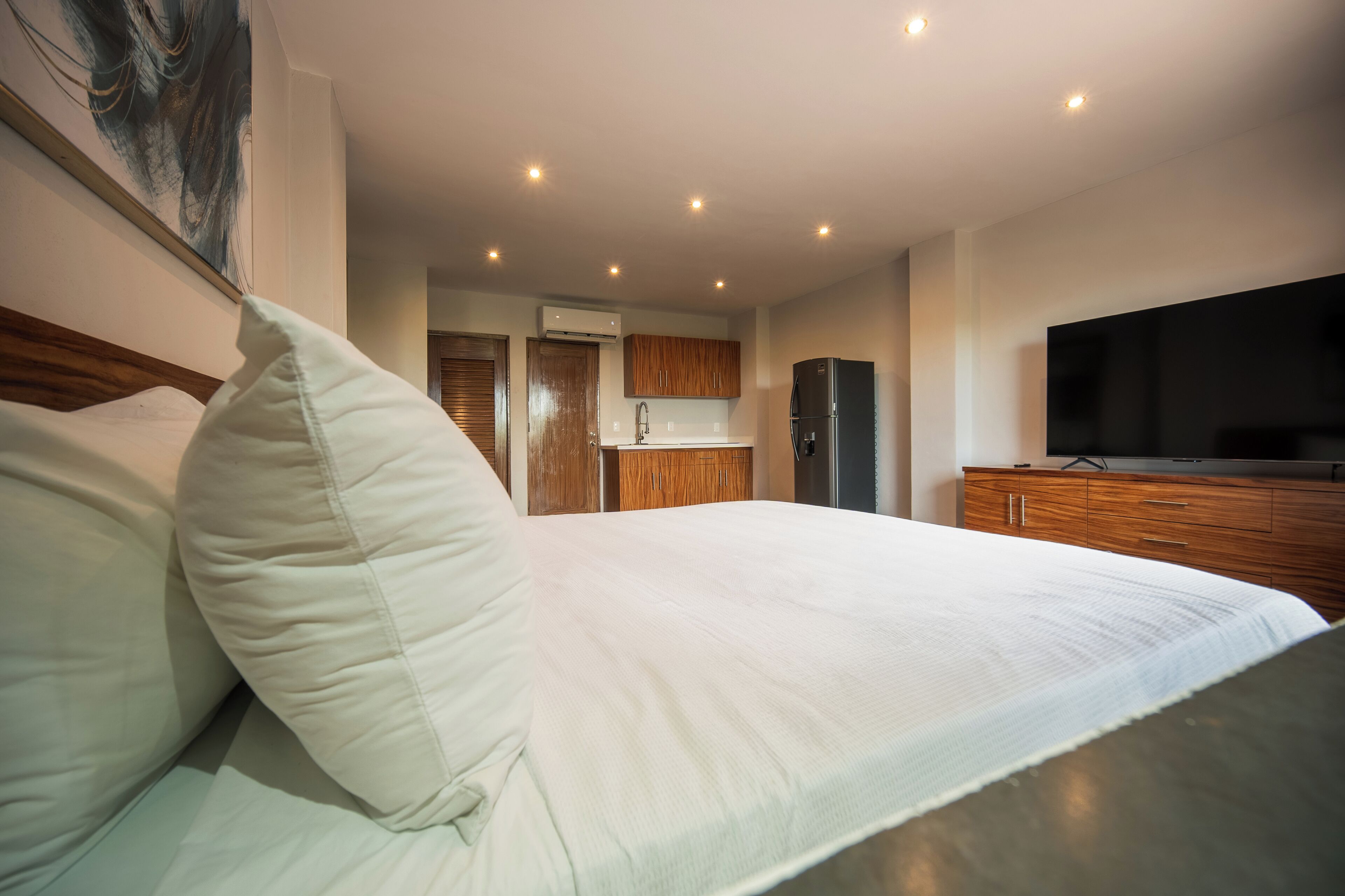 A modern hotel room with a large bed, wooden cabinets, a flat-screen TV, and a kitchen area in the background.