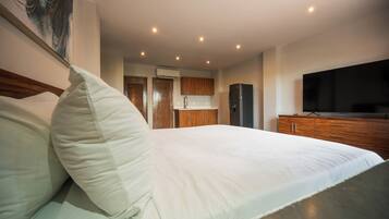 A modern hotel room with a large bed, wooden cabinets, a flat-screen TV, and a kitchen area in the background.