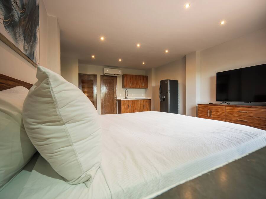 A modern hotel room with a large bed, wooden cabinets, a flat-screen TV, and a kitchen area in the background.
