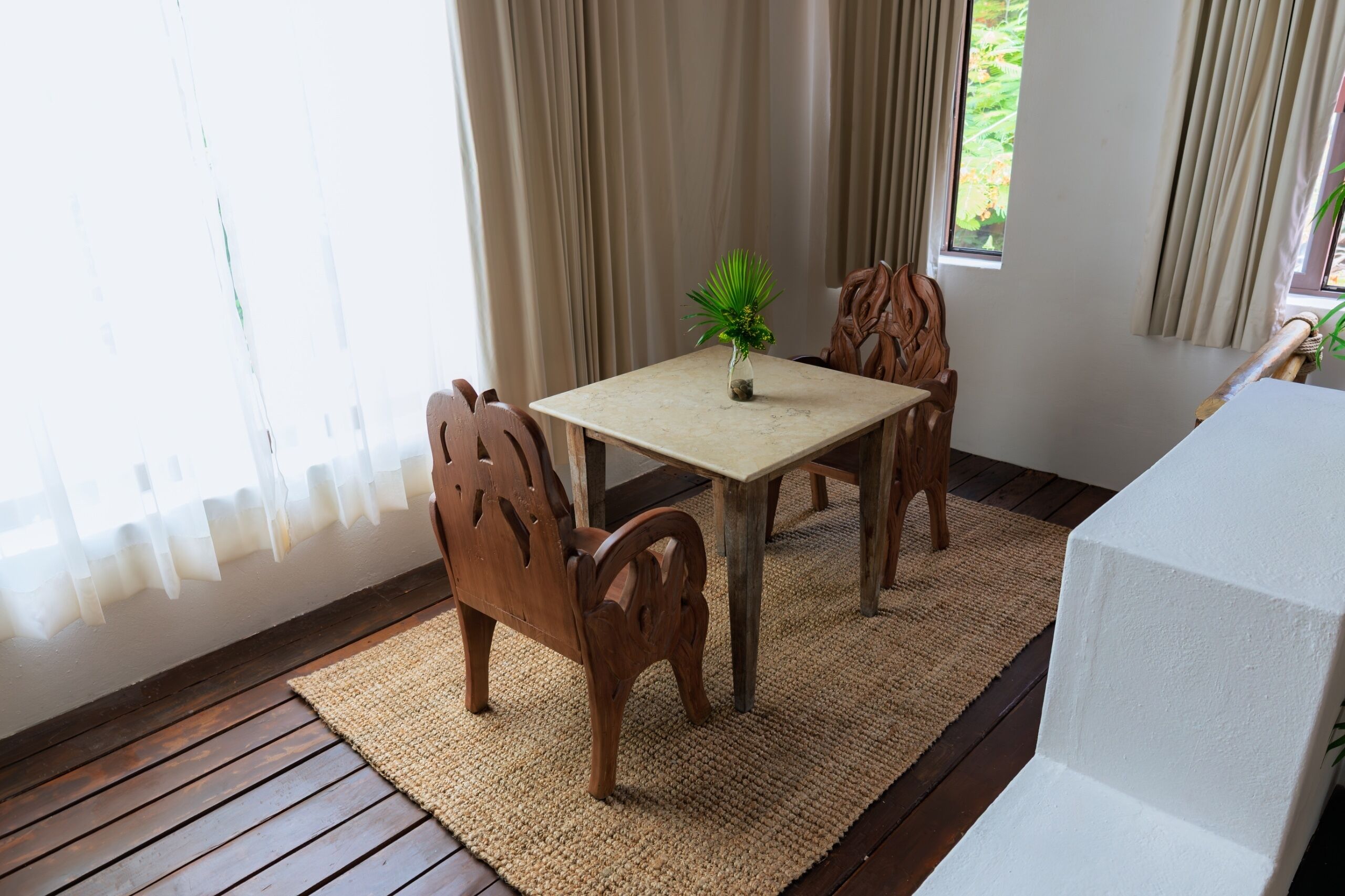 A wooden dining table with chairs, a small potted plant, and a view of the outdoors through a window with curtains.