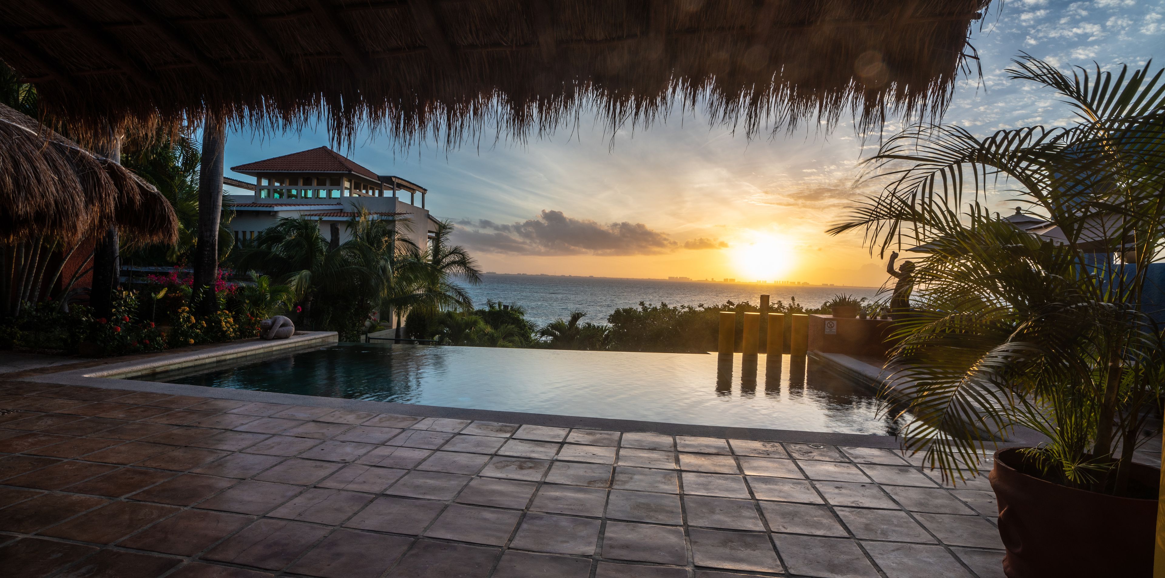 Un área de piscina con vista al atardecer sobre el océano, una estructura con techo de paja y plantas tropicales.