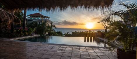 Un área de piscina con vista al atardecer sobre el océano, una estructura con techo de paja y plantas tropicales.